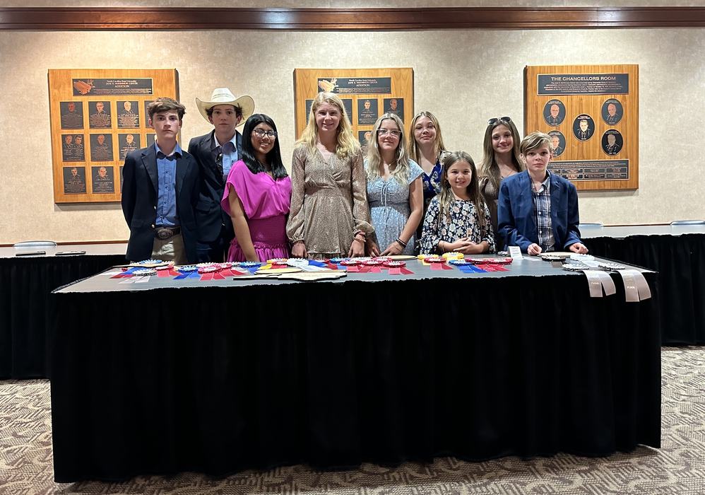 Ten young people standing behind a table covered with award ribbons in a conference room