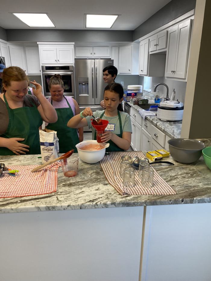 Group of children in a kitchen; girl labeled "Shelby" pours red mixture through a funnel into a jar