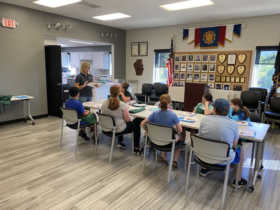 Woman instructing a group of children seated around tables in a meeting room with an American flag