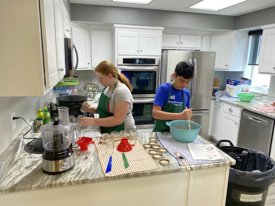 Two children in aprons stirring batter and filling jars on a kitchen island.
