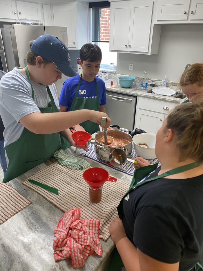 Group of children in aprons ladling tomato sauce into jars on a kitchen counter