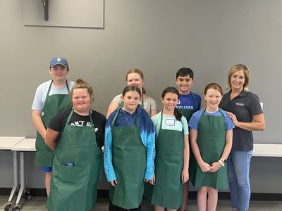 Group of children and an adult wearing green aprons in a classroom; name tag "Shelby"
