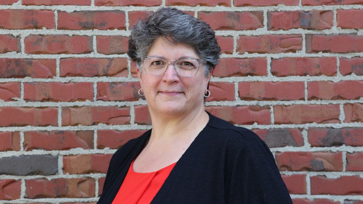 Person with short gray hair and glasses wearing a red top and black cardigan standing by a brick wall