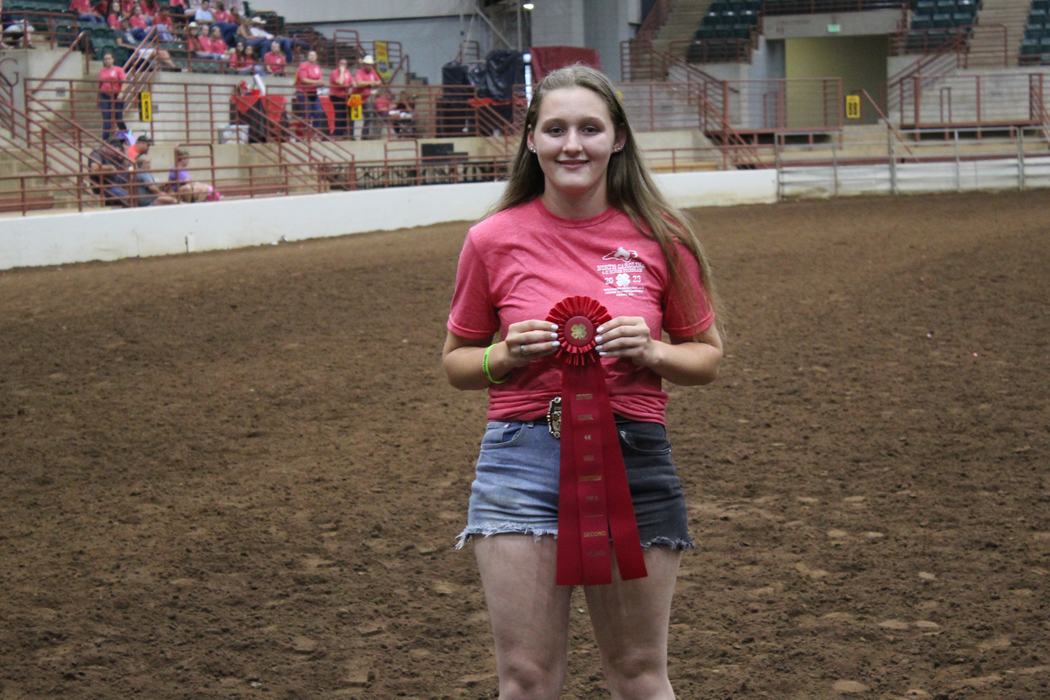 Young woman holding red 4‑H ribbon in an indoor dirt arena