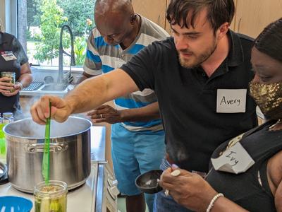 Man with "Avery" name tag stirring large pot while woman with "Ivy" name tag jars food