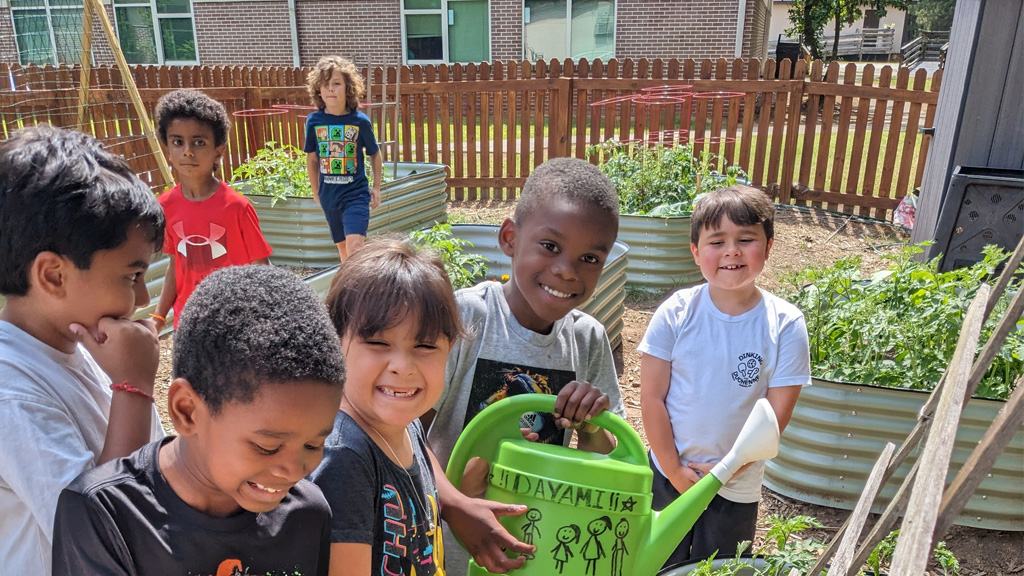 Group of children in a raised-bed garden, boy holding green watering can with handwritten text