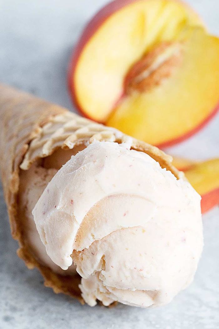 Peach ice cream scoop in a waffle cone with sliced peach halves in background