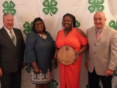 Four individuals pictured with award recipient in front of a green and white 4-H drop.