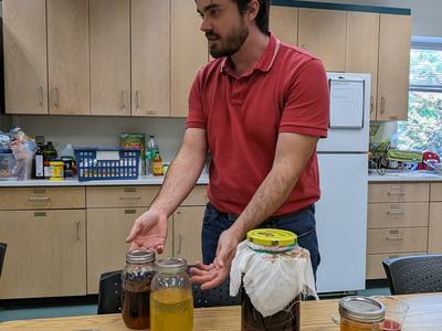 Person in red shirt demonstrating jars of kombucha and a SCOBY on a table in a kitchen