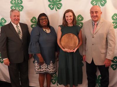 Four people standing before 4‑H clover backdrop; center woman holds wooden award plaque