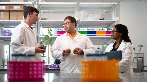 Three NC State Extension interns wearing white lab coats talk in a scientific lab setting.