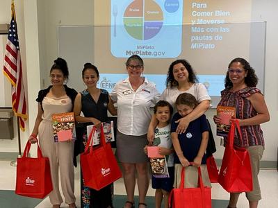 Six adults and two children holding red EFNEP bags and cookbooks before a MiPlato (MyPlate.gov) slide.