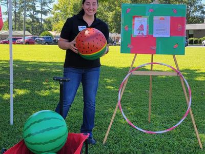 Person holding a watermelon-patterned ball beside a wagon of balls and a hoop game board outdoors