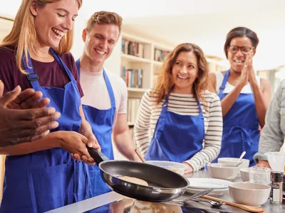 Woman flipping pancake in frying pan while a group in blue aprons watches
