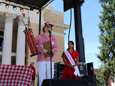 Person holding large trophy and speaking at microphone; woman beside wearing sash "NC Apple Ambassador"