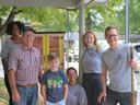 Six people posing outdoors beside a small painted free library under a porch.