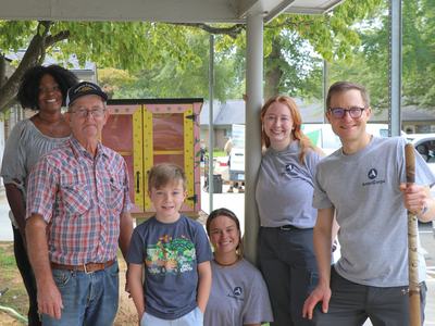 Six people posing outdoors beside a small painted free library under a porch.