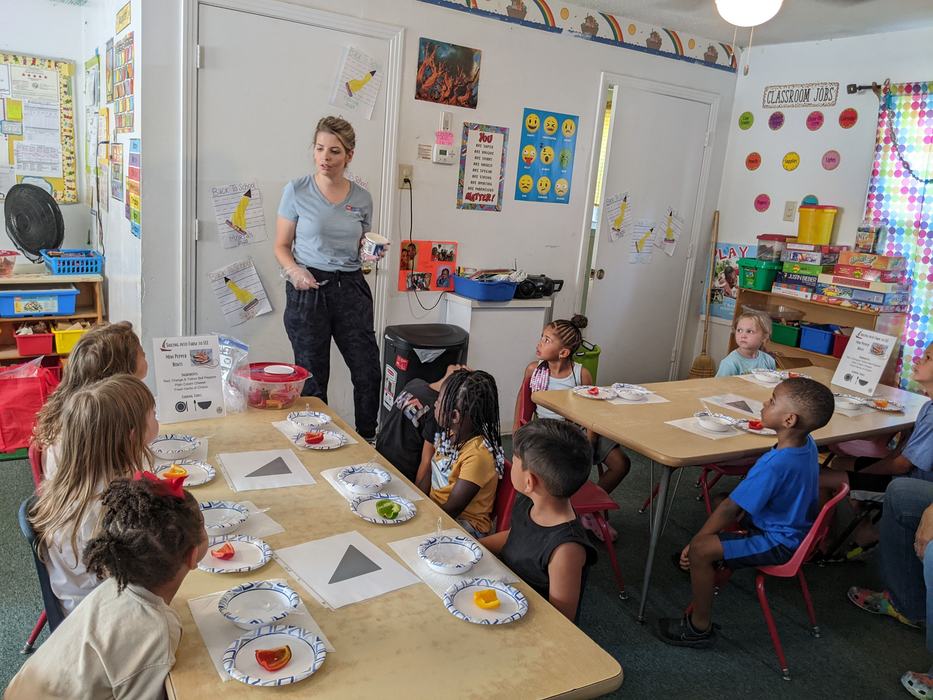 Teacher standing in classroom instructing seated children at tables with snack plates
