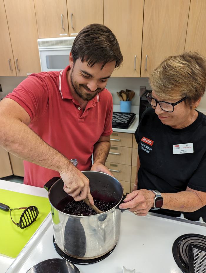Two people work on stirring preserves in a large pot