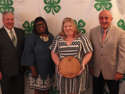 man in suit, woman in dress, woman in dress holding wooden plaque, man in tan jacket