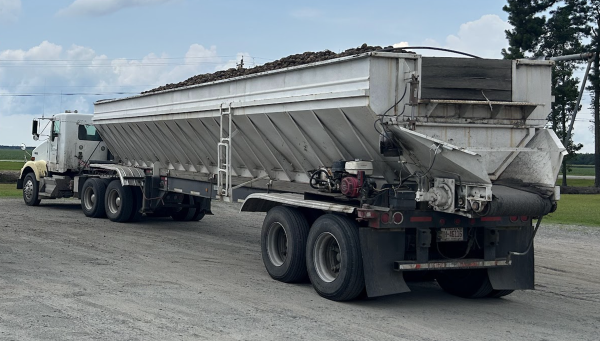 White semi truck parked on gravel hauling a long white hopper trailer loaded with bulk loose cargo
