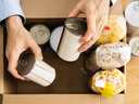 Hands placing canned goods, pasta, and oats into a cardboard food donation box