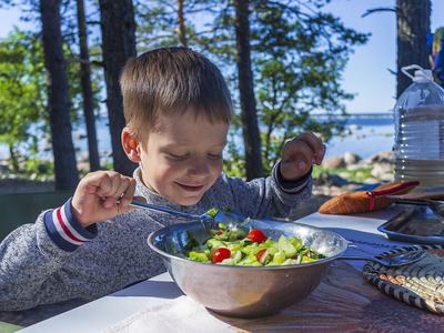 Young child scooping cucumber and tomato salad from a metal bowl at an outdoor lakeside table