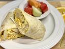 Breakfast burrito cut in half on a white plate with a small bowl of mixed fruit