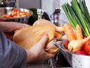 Hands holding a raw whole chicken under running faucet with vegetables nearby