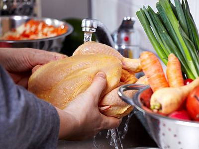 Hands holding a raw whole chicken under running faucet with vegetables nearby