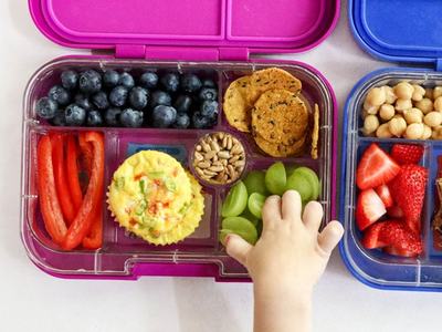 Child's hand reaching into compartmented lunchbox with grapes, blueberries, mini frittata, crackers