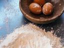 Three brown eggs with flour in a wooden bowl beside a mound of flour on a dark surface