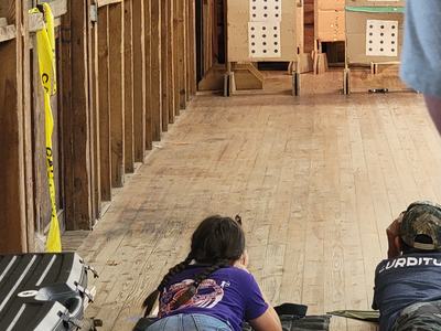 Two children lying prone on mats aiming at numbered target boards (1 and 2) in indoor range