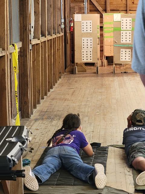 Two children lying prone on mats aiming at numbered target boards (1 and 2) in indoor range