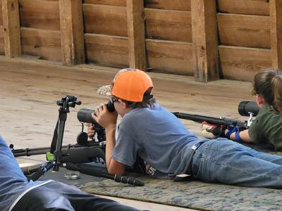 Child in orange cap lying prone aiming a scoped rifle on indoor wooden floor