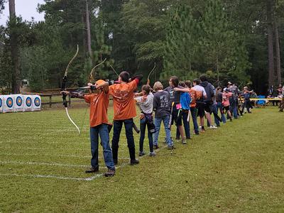 Youth archers standing in a line aiming bows at targets on a grassy outdoor range