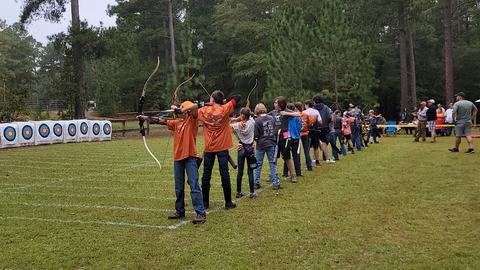 Youth archers standing in a line aiming bows at targets on a grassy outdoor range