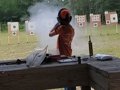 Shooter with ear protection firing a shotgun at outdoor paper targets, muzzle smoke visible