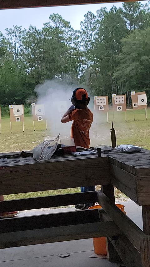 Shooter with ear protection firing a shotgun at outdoor paper targets, muzzle smoke visible