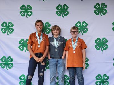 Three youth wearing medals standing in front of a repeated 4‑H clover backdrop