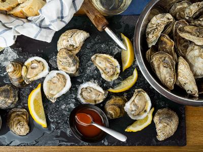 Dark marble serving platter with shucked oysters with sliced lemons and a sppon of cocktail sauce on a table next to a bucket of unshucked oysters and a basket with a tea towel and bread in it.