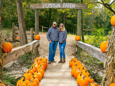 Couple standing under "PUMPKIN PATCH" sign on wooden bridge lined with pumpkins