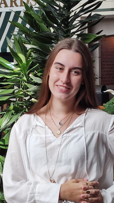 Sarah Beth has long brown hair and smiles at the camera wearing a white shirt. 