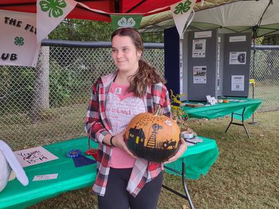 Young person holding a painted pumpkin under a red tent with 4‑H clover banners