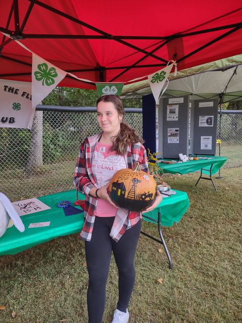 Young person holding a painted pumpkin under a red tent with 4‑H clover banners