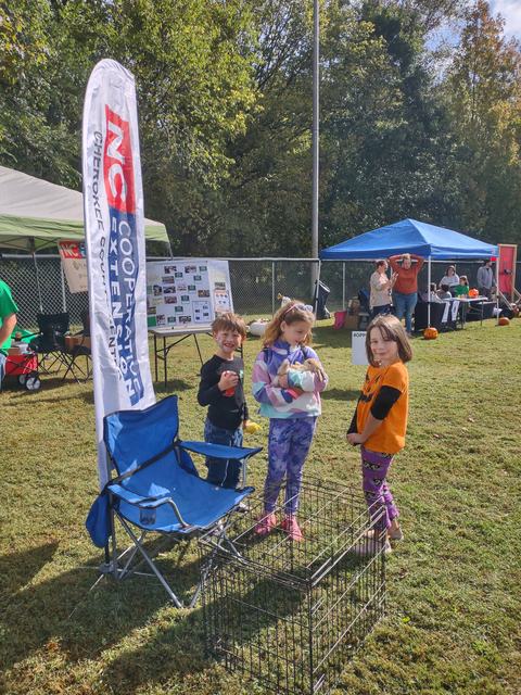Three children at an outdoor booth, one holding a small rabbit; banner reads "NC Cooperative Extension"