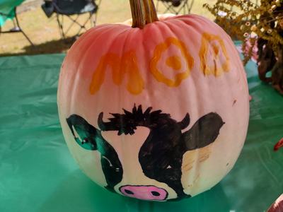White pumpkin painted with a cow face and "MOO" on a table at an outdoor fair