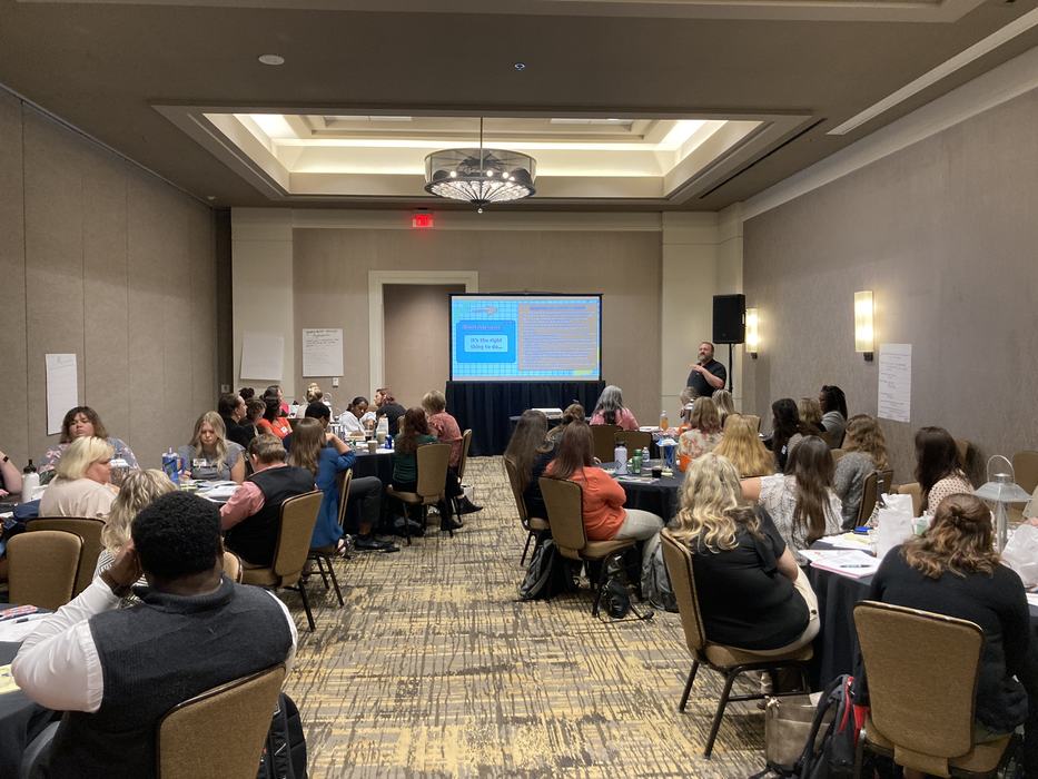 4-H Extension agents and program assistants sit at round tables in a hotel conference room. A 4-H specialist leads the discussion with a PowerPoint presentation at the front of the room.
