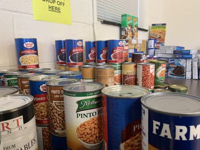 Canned beans and boxed food stacked on a table with a yellow "DROP OFF HERE" sign