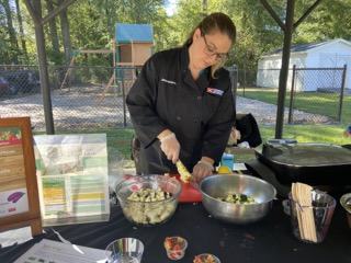 Person wearing dark jacket preparing salad at outdoor table with bowls and ingredients.